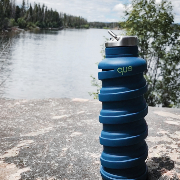Collapsible Water Bottle in Blue, shown on rock at edge of lake with trees in the distance.