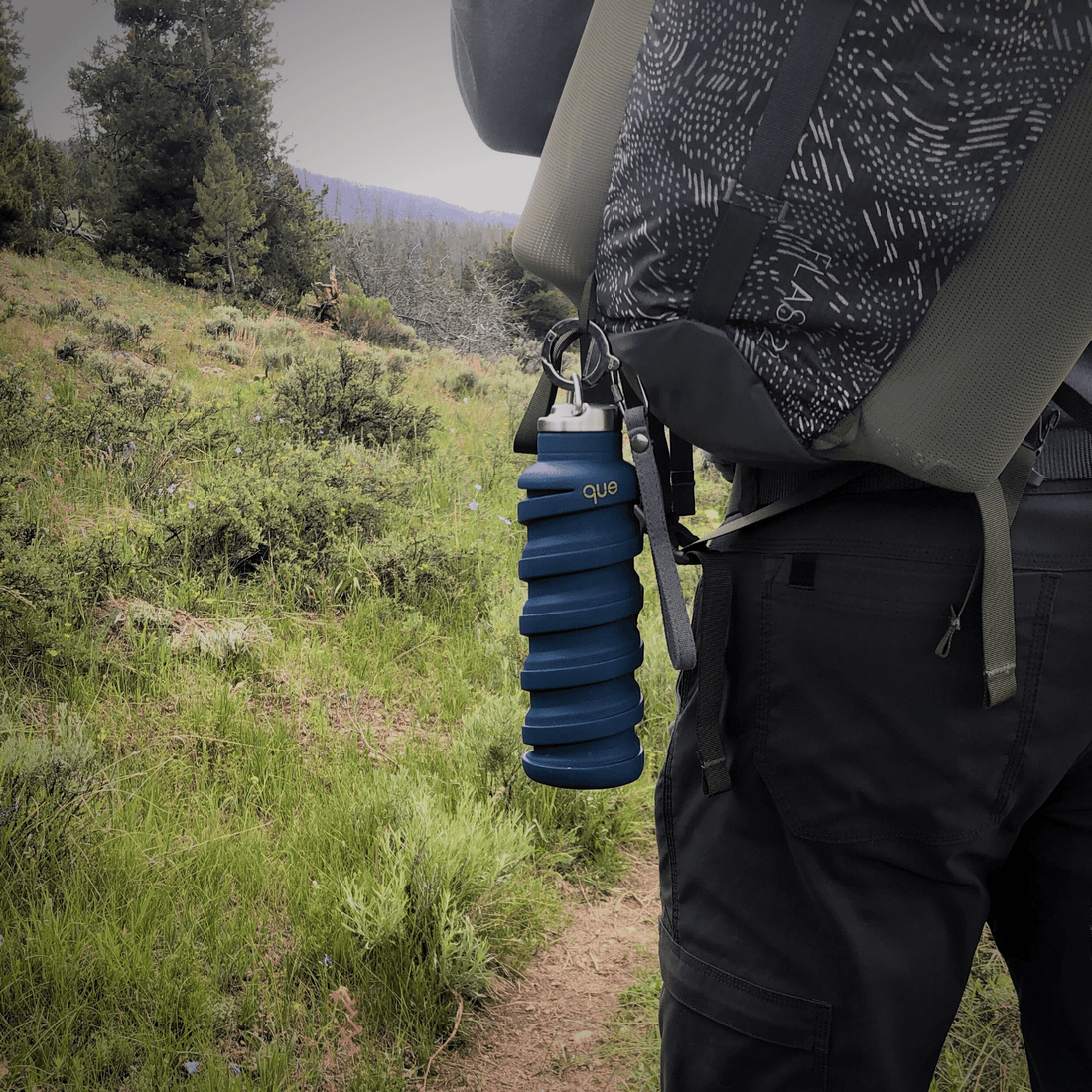 Que Bottle, Shown Expanded with Accessory Combo (Carabiner Bottle Opener, Bottle Cap with Loop, and Carry Strap) Sold Separately. Blue Que Bottle Hanging from Back Pack on Mountain Trail.