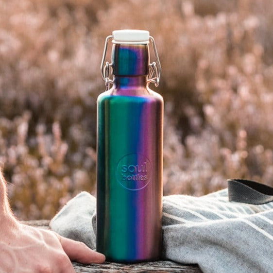 Soulbottles Utopia Stainless Steel Water Bottle - 20 ounce. Rainbow Colored and Plastic Free, Shown on Table In Field with Man's Hand and Sweatshirt.