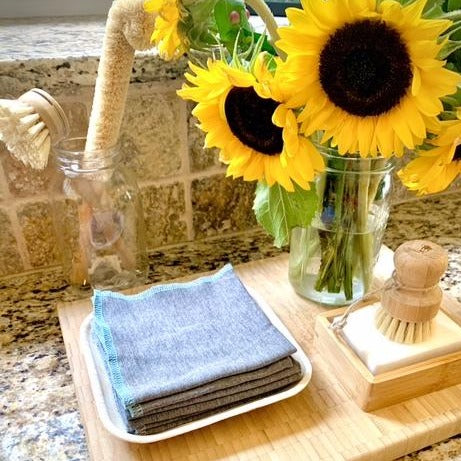 Reusable All Purpose Bamboo Eco Cloths With Recycled Thread Trim In Gray, Turquoise, And Blue Colors On Kitchen Counter With Cutting Board, Other Cleaning Items, And Jar Of Sunflowers.