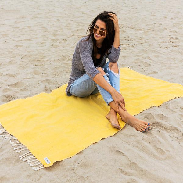 Sand Cloud Turkish Cotton Beach Travel Towel On Sand With Woman Relaxing And Smiling On Top Of Towel.