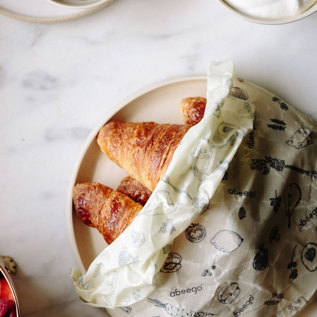 Abeego Beeswax Food Wrap. Large Size Shown Wrapped Around Plate with Croissants on Countertop.