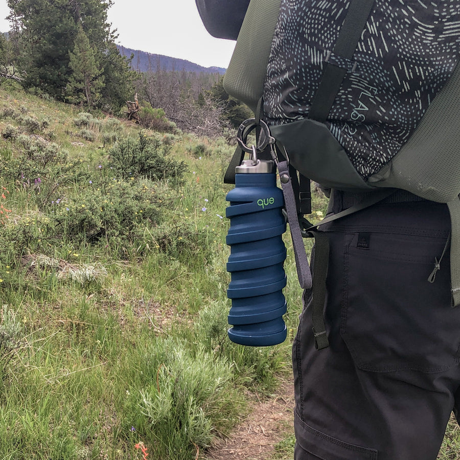 Que Bottle In Midnight Blue Hooked To Backpack With Bottle Cap With Loop And Accessory Combo Keychain and Strap. Mountain Trail In Background.