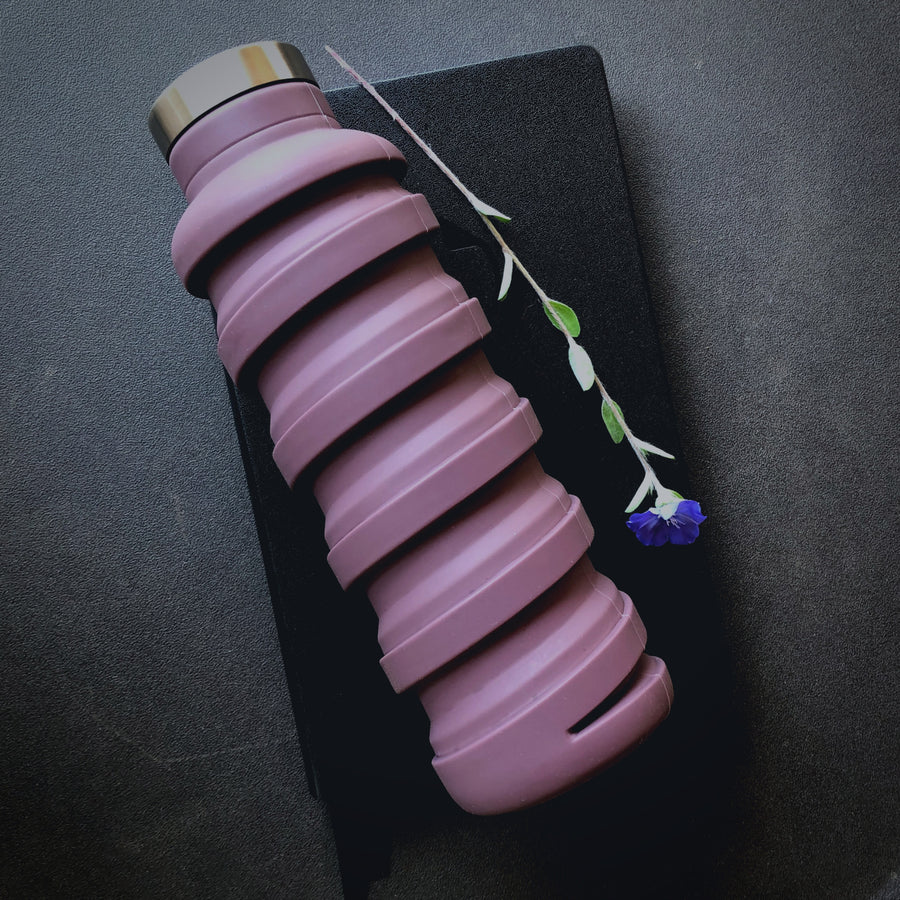Que Bottle In Frozen Plum Color With Cap. Shown Expanded For Use. Set On Notebook With Flower.
